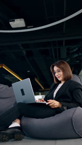 comfortably sitting young woman in a bean bag chair with laptop in her hands. low angle view on the lady freelancing and working remotely. vertical video.