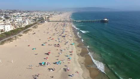 Vista-Lateral-Al-Mediodía-De-La-Playa-De-Manhattan-Llena-De-Bañistas-Nadando-Y-Surfeando-En-California-Usa---Toma-Aérea-Panorámica-Hacia-Adelante