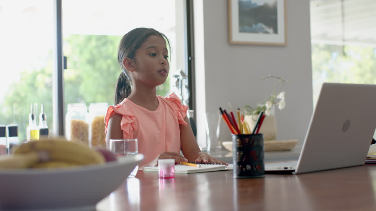Happy biracial girl having elementary school class on laptop in kitchen raising hand, slow motion