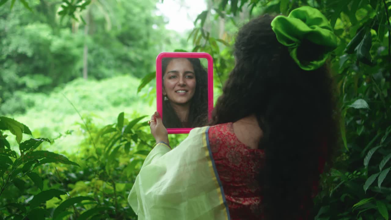 Curly-haired woman smiles at her reflection in a hand mirror in nature setting