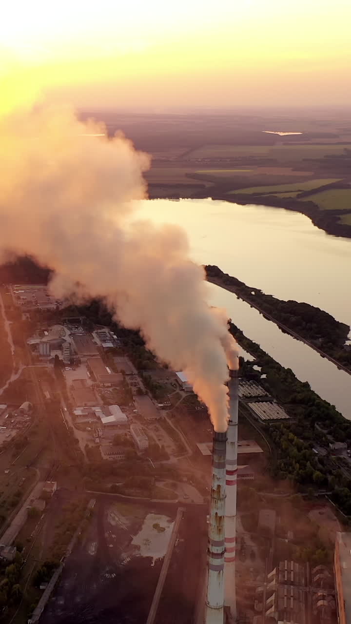 Aerial view of a factory among nature. Industrial pipes pollute the atmosphere with smoke at sunset. Beautiful river next to the huge metallurgy plant. Vertical video