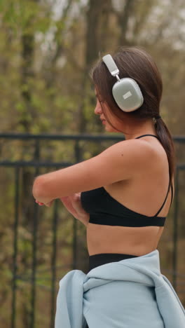 lady standing confidently under bright atmosphere, checking time on her wristwatch while listening to music with headphones, enjoying outdoor activity in a peaceful and vibrant park setting