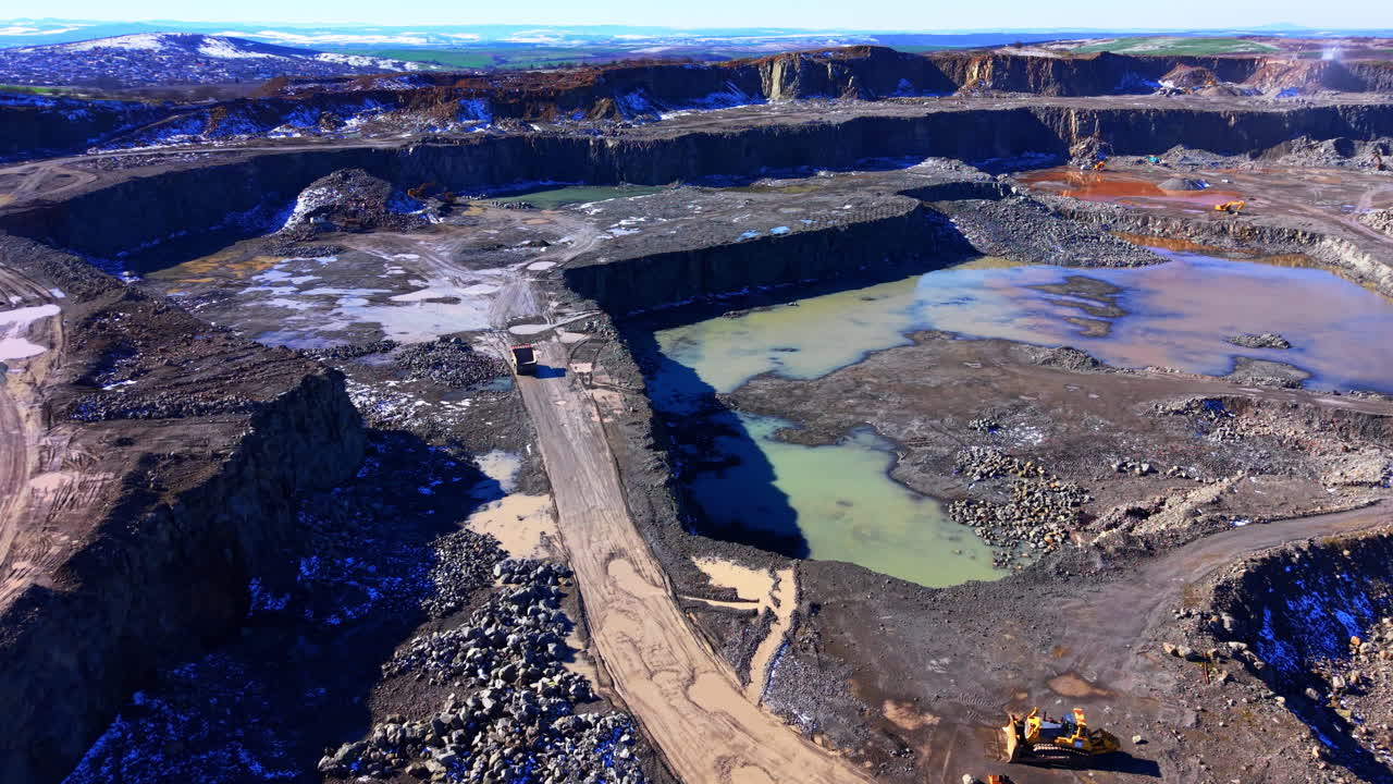 Panoramic view of a large stone quarry with layered rock walls, water pools, and heavy machinery including a bulldozer and trucks working on the site