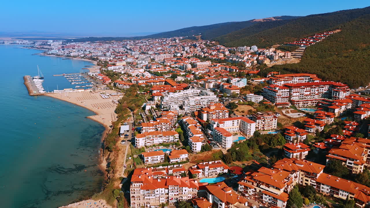 Resort balneare con vista panoramica. Veduta aerea di una vivace località costiera con edifici colorati, un porto turistico e un lungomare suggestivo sotto un cielo sereno.