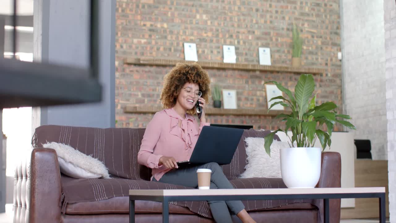 Happy biracial casual businesswoman using laptop talking on phone on couch in office, slow motion