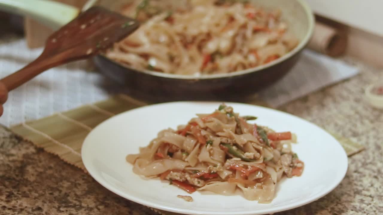 Serving stir-fried rice noodles on a white plate, with additional noodles in a pan nearby, depicting a warm kitchen setting and home cooking