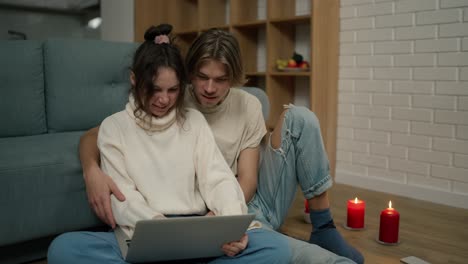 Young-adult-couple-using-laptop-computer-at-home-sitting-on-floor-around-candles