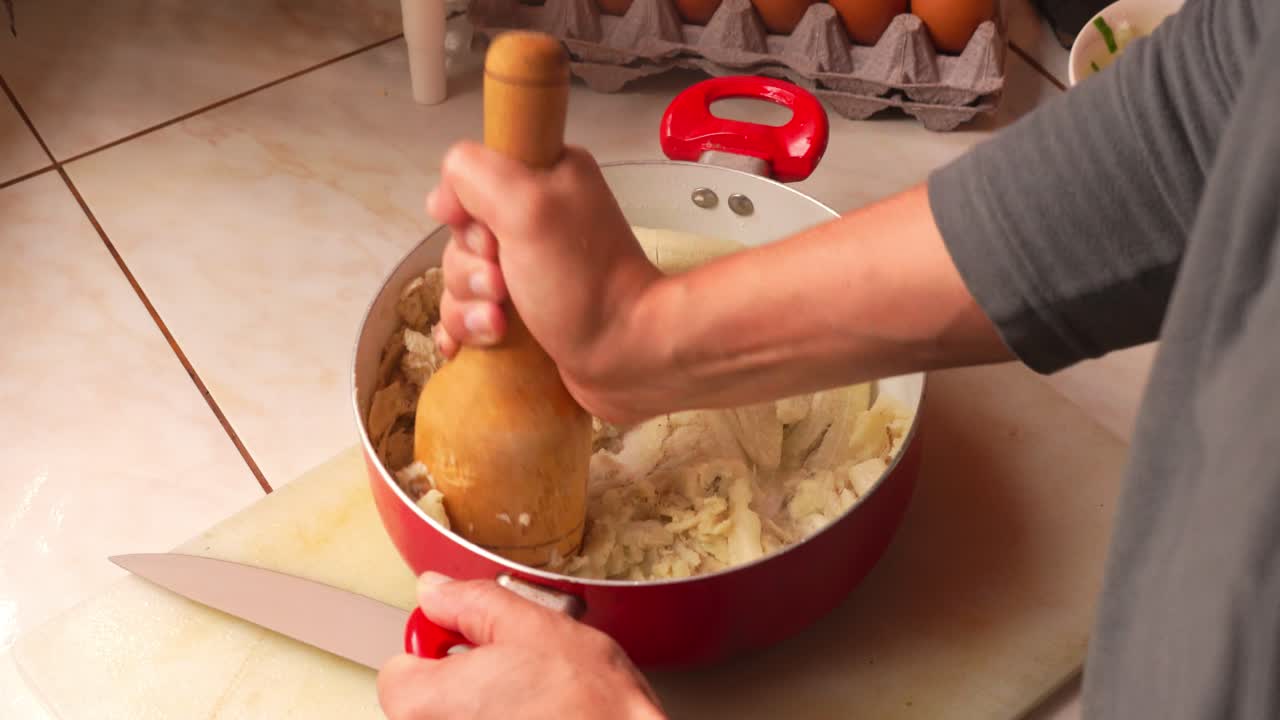 Close-up of a person mashing green plantains in a red pot using a wooden tool. Ideal for traditional food prep, recipes, or editorial use.
