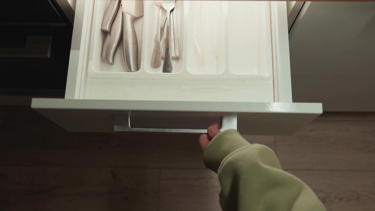 Una scena dall'alto di una cucina domestica mostra una mano con una manica verde che apre un cassetto di legno per rivelare un vassoio portaposate bianco con forchette, coltelli e scomparti in acciaio inossidabile, uno stoccaggio pulito e organizzato.