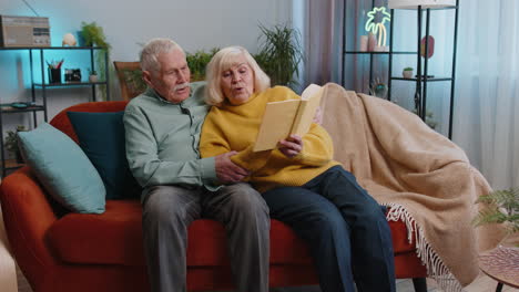 senior family couple man woman enjoying reading interesting book, talking, laughing at home on sofa