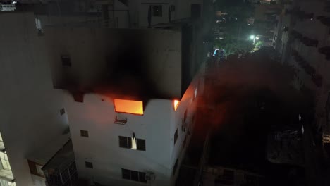 smoke leaving burning house at night. flames inside room of apartment in city. aerial view.