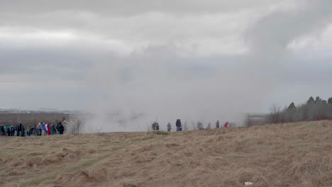 static, wide shot, of lots of people standing in front of the erupting geisir, on a cloudy, autumn day, in geysir, iceland