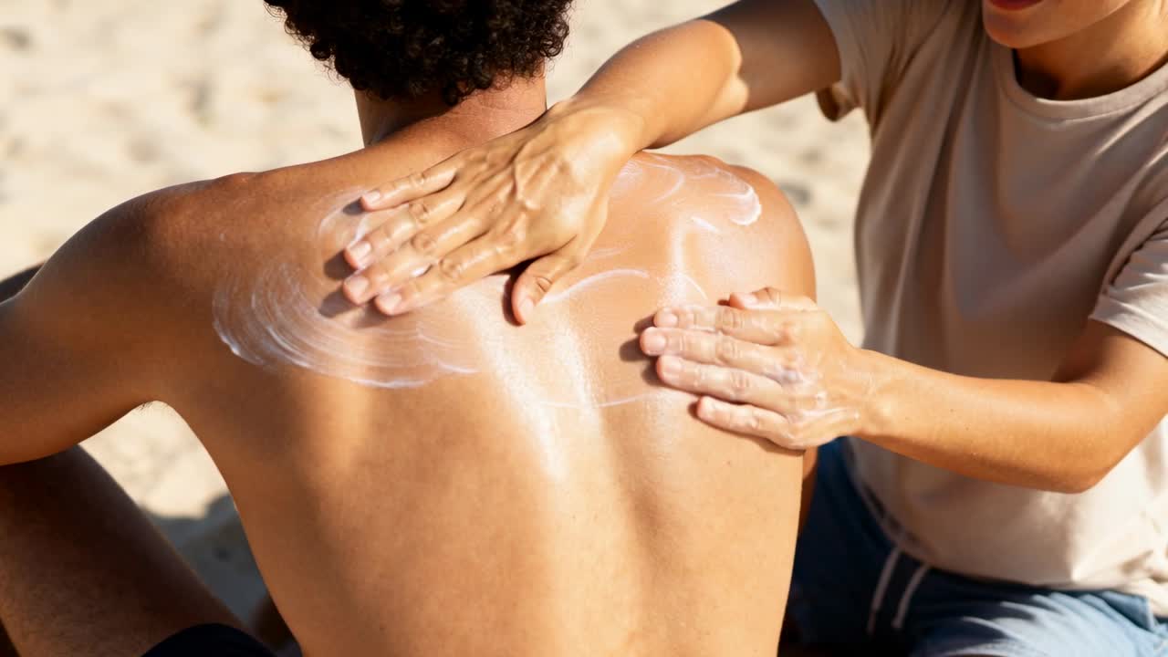 Placing two dots, partner rubbing sunscreen onto man in swim shorts on sand, protecting from sun