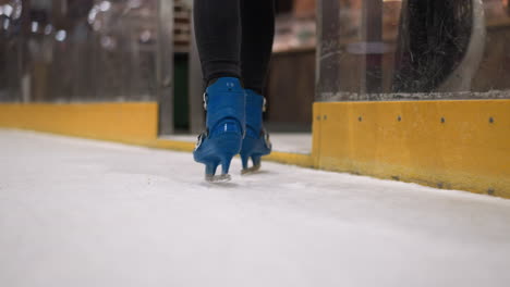 close-up of someone skating on an ice rink wearing blue skates and black trousers, walking out of the rink