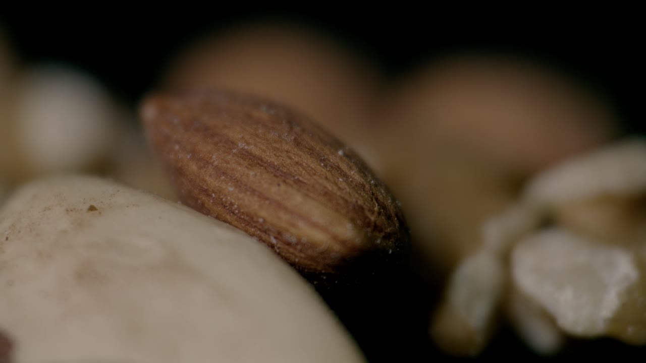 Primo piano ravvicinato di un assortimento di frutta secca, scattato con illuminazione drammatica per mettere in risalto texture e dettagli. Il focus è su una singola mandorla con frutta secca sfocata sullo sfondo, per evocare un’atmosfera rustica