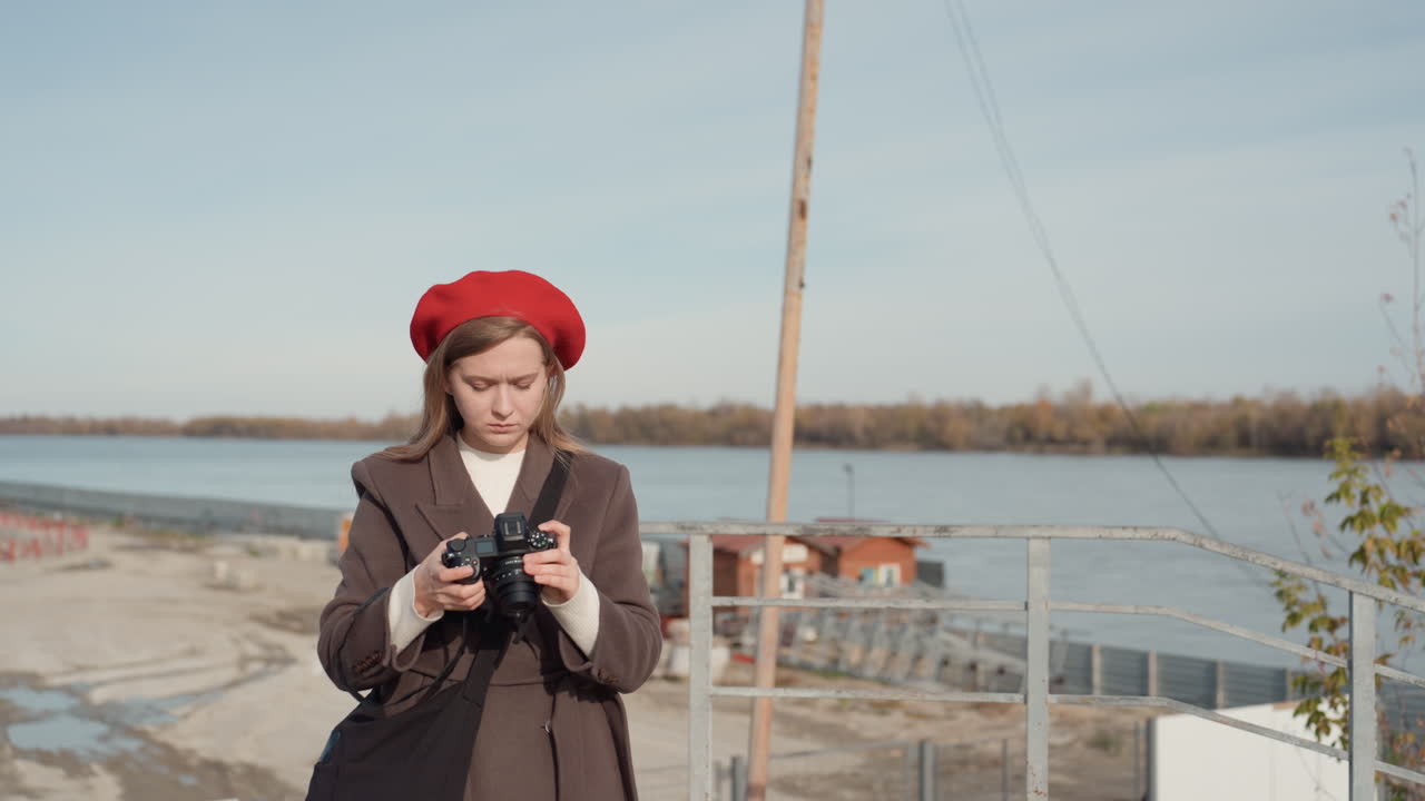 Молодая женщина исследует индустриальную набережную, в красном берете и длинном пальто, настраивает объектив и кадрирует снимки, проверяет экспозицию при ярком дневном свете, доки и склады на заднем плане, современный инди-стиль.