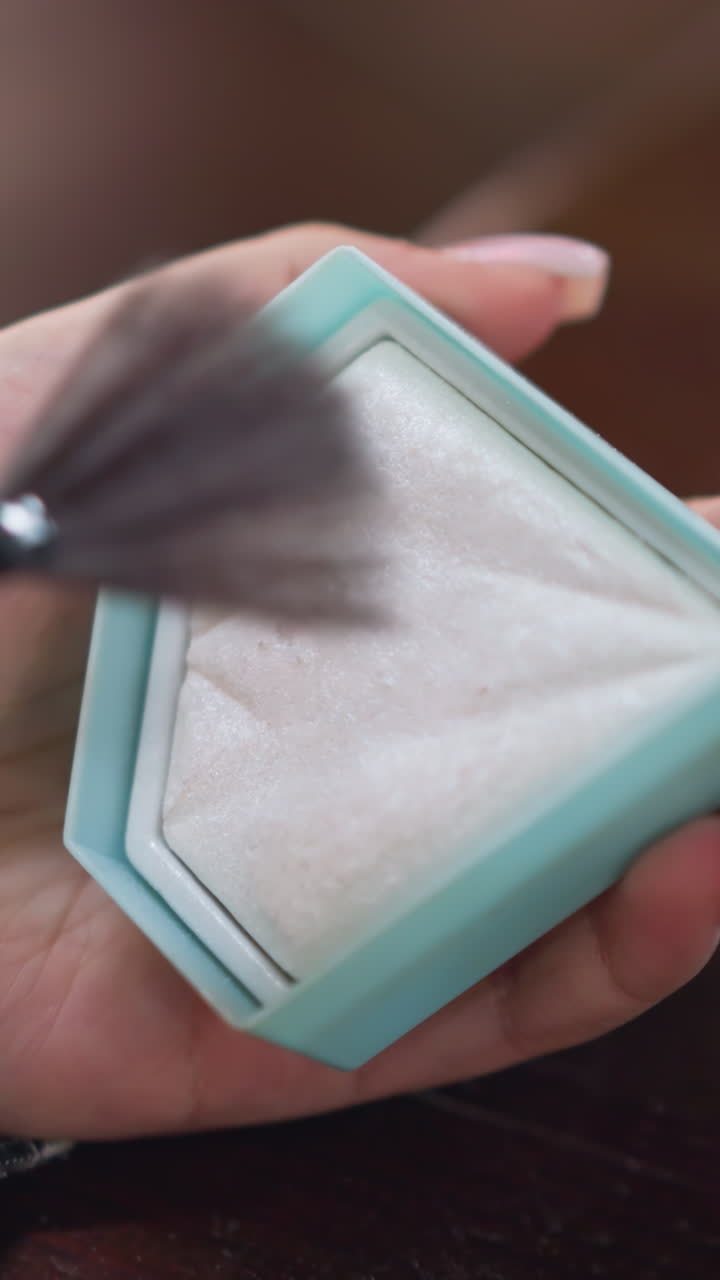 Primer plano de una mano sosteniendo una brocha abanico compacta de color turquesa, que aplica iluminador nacarado sobre una lujosa polvera blanca; las uñas cuidadas y el movimiento suave crean un brillo sutil y un acabado de lujo, enfatizado por la iluminación de estudio