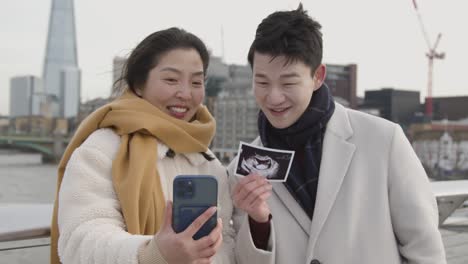 영국 런던에 있는 임신한 젊은 아시아 부부가 가족에게 신생아의 초음파 사진을 보여주는 전화로 집에 화상 통화를 하고 있습니다.