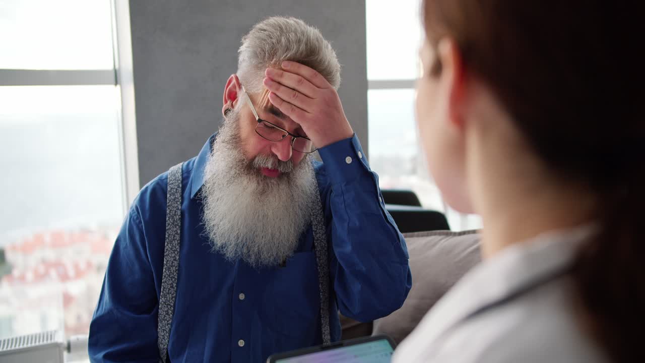 Over the shoulder a man with gray hair and glasses with a lush beard in a blue shirt complains to a woman doctor about his problems and headaches during a home examination in a modern apartment