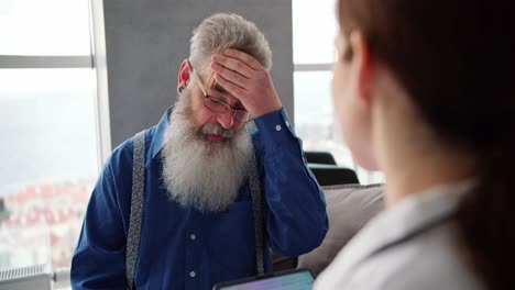 Por-Encima-Del-Hombro,-Un-Hombre-De-Pelo-Gris-Y-Gafas-Con-Una-Barba-Exuberante-Y-Una-Camisa-Azul-Se-Queja-Con-Una-Médica-Sobre-Sus-Problemas-Y-Dolores-De-Cabeza-Durante-Un-Examen-Domiciliario-En-Un-Apartamento-Moderno.