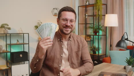 young businessman freelancer holding fan of cash and showing thumbs up sitting at home office table