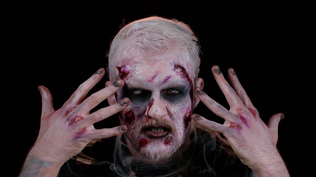 aterrador zombie herido no muerto tipo haciendo caras, mira a la cámara escondiéndose a través de las manos sonríe terriblemente