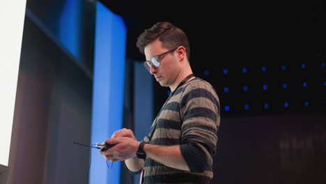 technician wearing striped sweater and glasses inspects wireless microphone transmitter cables using audio device inside illuminated television studio with blue backdrop during live broadcast prep