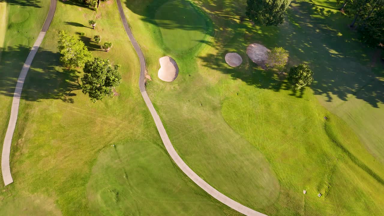 Drone footage captures a lush golf course with vibrant greens, sand bunkers, and winding paths under bright sunlight