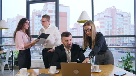 female worker stands near the boss sitting in front of laptop. lady speaks and points at computer. chief in a hurry pointing at his watch. other employees at backdrop.
