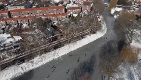 top down urban aerial winter scene with people ice skating along the curved frozen canal going through the dutch city of zutphen with shadows of barren trees and former water tower in the background