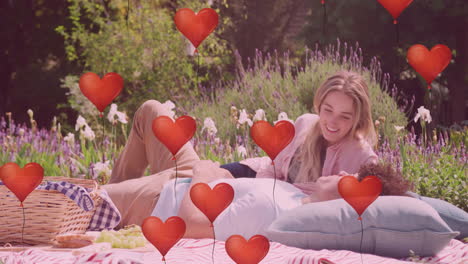 Animation-of-red-heart-balloons-over-happy-diverse-couple-relaxing-at-picnic-in-sunny-garden