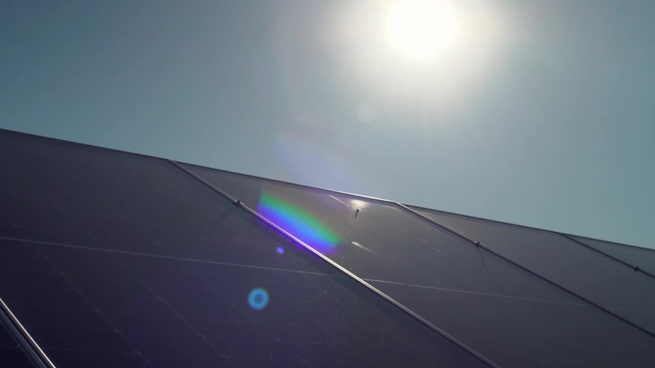 Sole luminoso con un vivace flare dell'obiettivo sulla superficie di vetro scuro di un impianto fotovoltaico a pannelli solari sul tetto. Ampia vista dal basso.