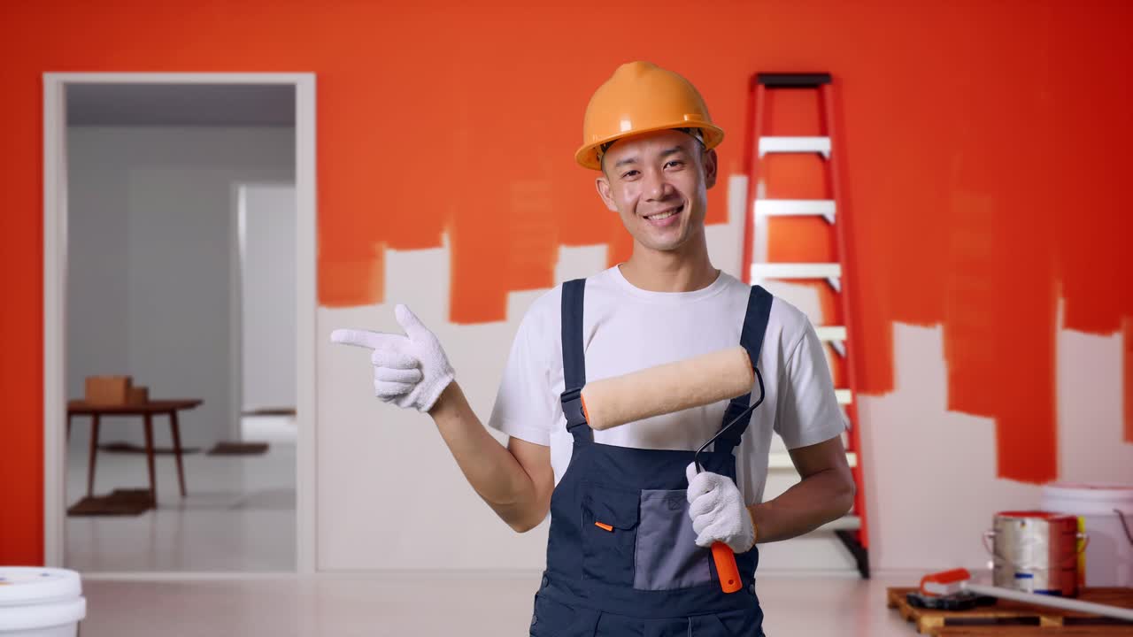 Asian Man Painter Wearing Safety Helmet Smiling And Pointing To Side While Standing With Painting Wall In Room