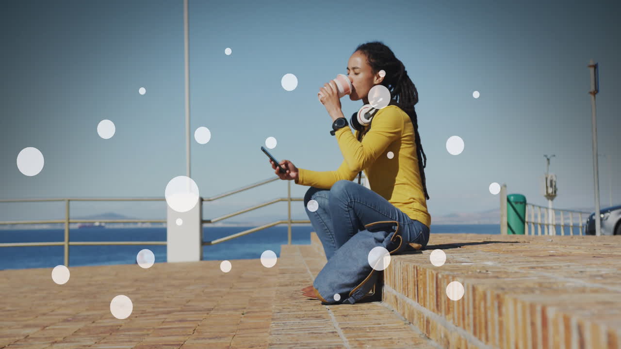 Punti bianchi su donna afroamericana che beve caffè e usa lo smartphone sul lungomare