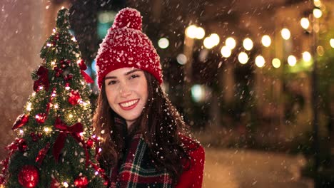 vista de cerca de una mujer caucásica con abrigo rojo sosteniendo un árbol de navidad y sonriendo a la cámara en la calle mientras nieva en navidad