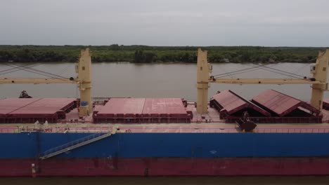 Aerial-close-up-of-old-cargo-ship-transporting-shipment-and-goods-cruising-on-river-with-forest-in-backdrop---South-America,-Argentina