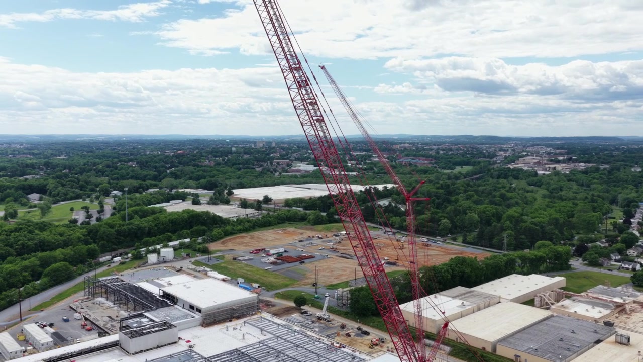 Towering red cranes rise above new data center site in Lancaster, with cleared land, staging buildings, and forest stretching toward distant horizon. Pennsylvania, USA. Aerial drone flyover shot
