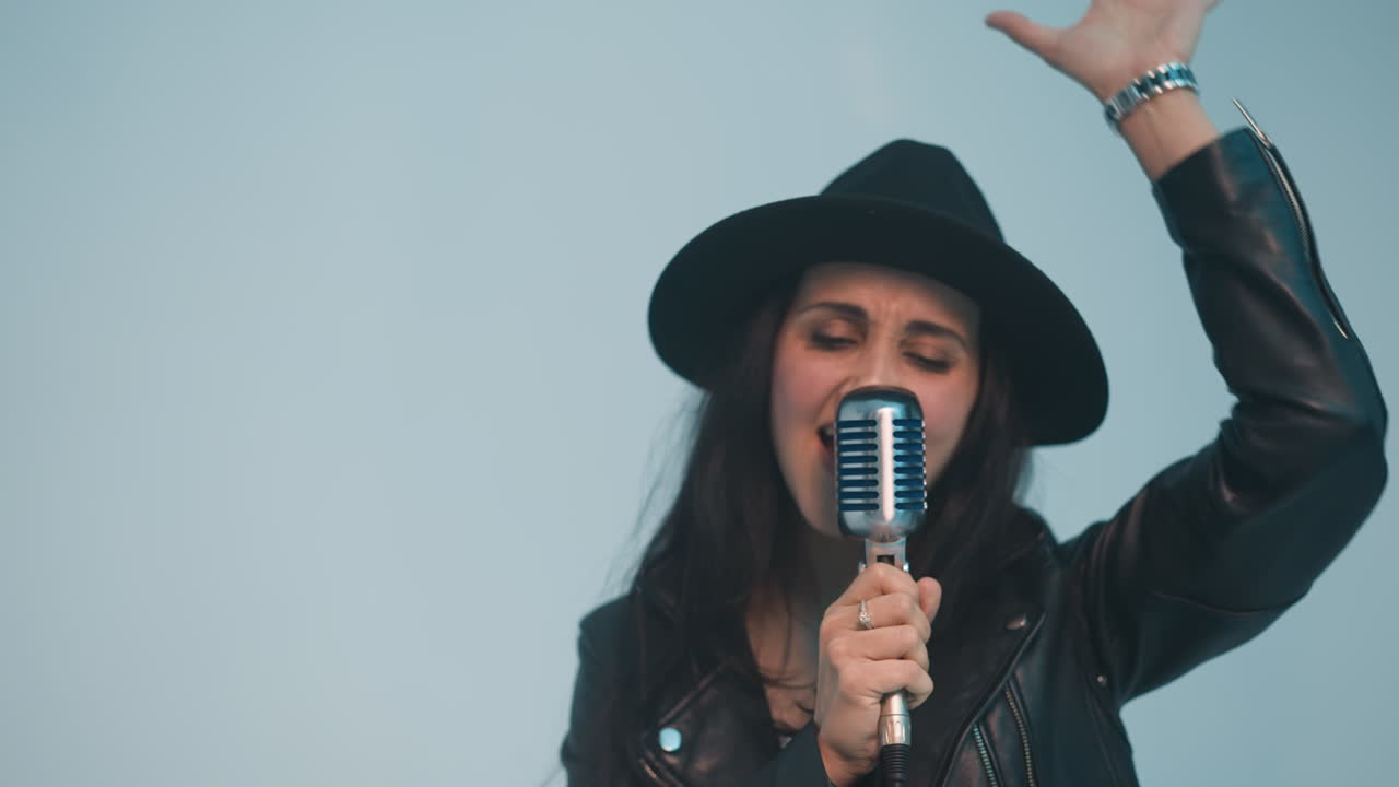 검은 모자와 가죽 재을 입은 열정적인 여성 가수가 한 손으로 빈티지 마이크를 들고 다른 손으로 부드러운 색 배경에 움직이고 있습니다.