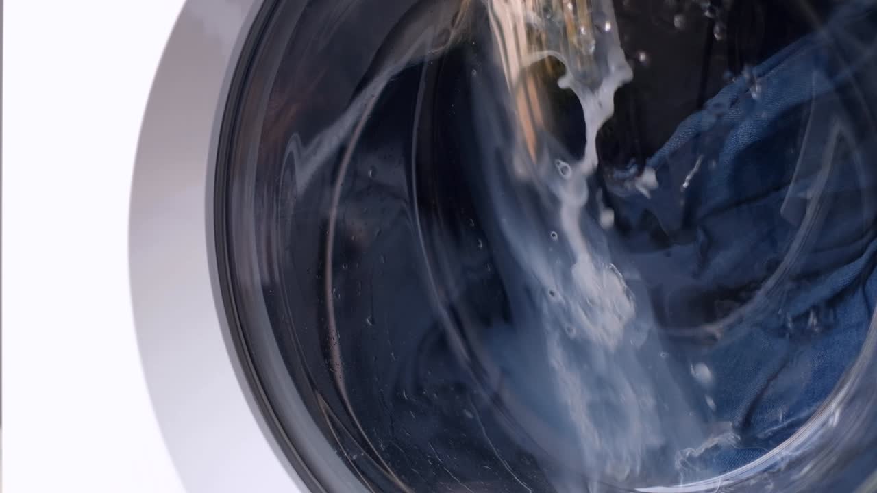 la ropa se lava en una lavadora automática blanca detrás de una puerta de vidrio, el tambor de metal gira en diferentes direcciones a su vez. lavado de ropa vaqueros. tarea 4k