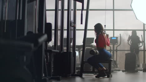 A-young-woman-in-blue-leggings-and-a-pink-top-performs-exercises-on-a-group-of-back-muscles.-Daily-training-in-the-gym-helps-to-maintain-a-healthy-body-and-spirit.