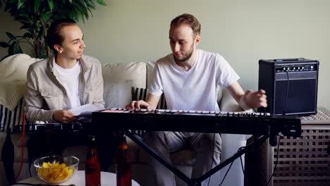 young bearded keyboarder is adjusting musical instrument then playing looking at music sheet and talking to his friend guitarist who is sitting on sofa in studio.