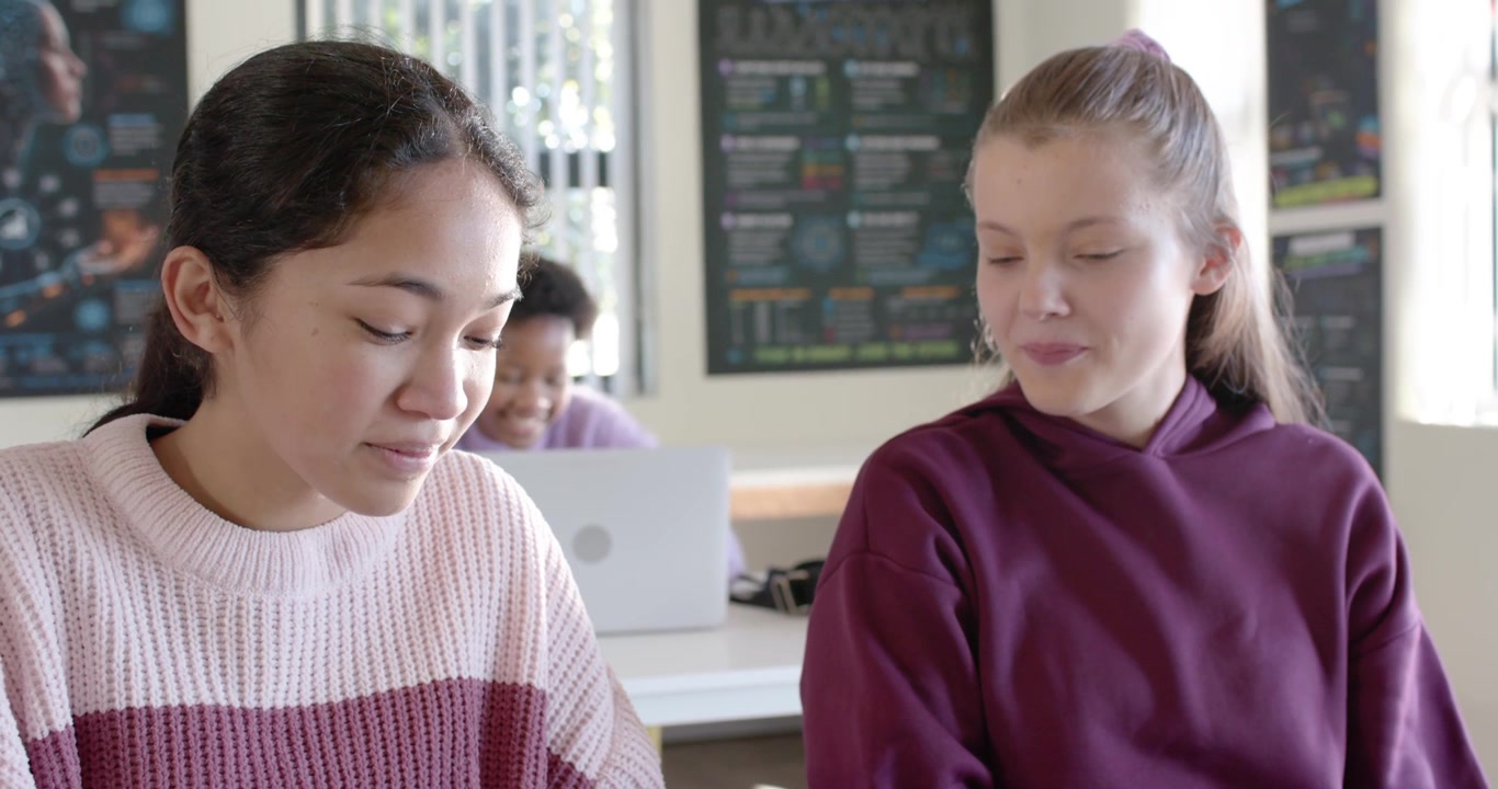 Two teenage girls at class table, left girl glancing, reaching phone, tapping, showing it by laptop