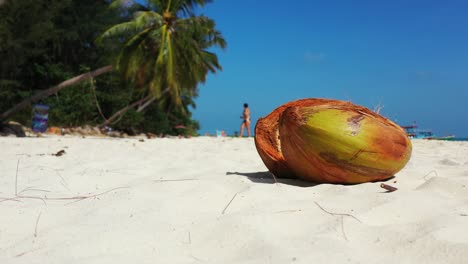 Nahaufnahme-Einer-Geknackten-Kokosnuss-Am-Sandstrand-Mit-Palmen-Und-Jungen-Mädchen,-Die-Im-Hintergrund-Auf-Dem-Sand-Spazieren-Gehen