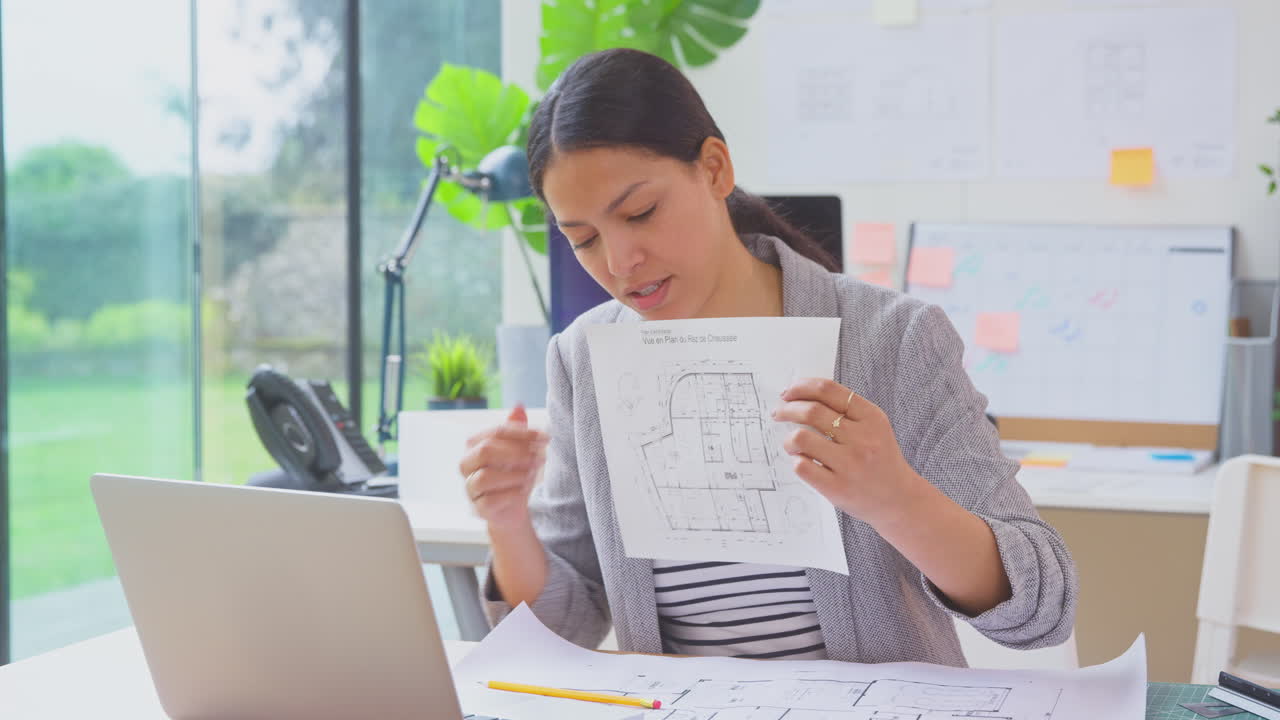 사무실에서 노트북을 가진 여성 건축가가 책상에 앉아서 비디오 통화로 새로운 건물의 계획을 보여줍니다.