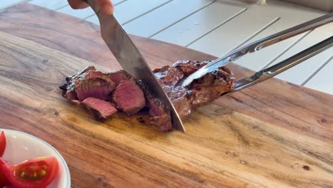 a hand uses a chef’s knife and tongs to slice a juicy, medium-rare wagyu steak on a wooden cutting board in bright, natural light