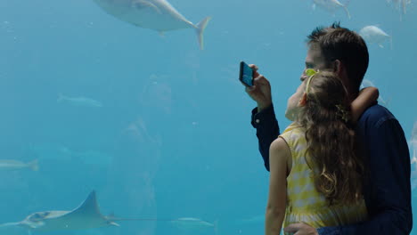 father with little girl at aquarium taking photos of fish using smartphone enjoying marine life swimming in tank having fun at oceanarium sharing on social media