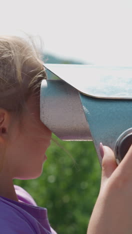 little girl with plait looks through large spyglass with attentive mother help on view point on sunny day close side view slow motion