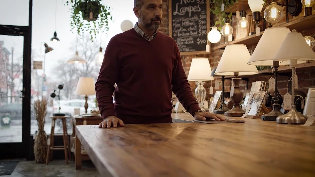 Un uomo pensieroso in un accogliente negozio di illuminazione, sotto la pioggia, circondato da lampade decorative e un'atmosfera calda che esalta l'ambiente invitante all'interno.