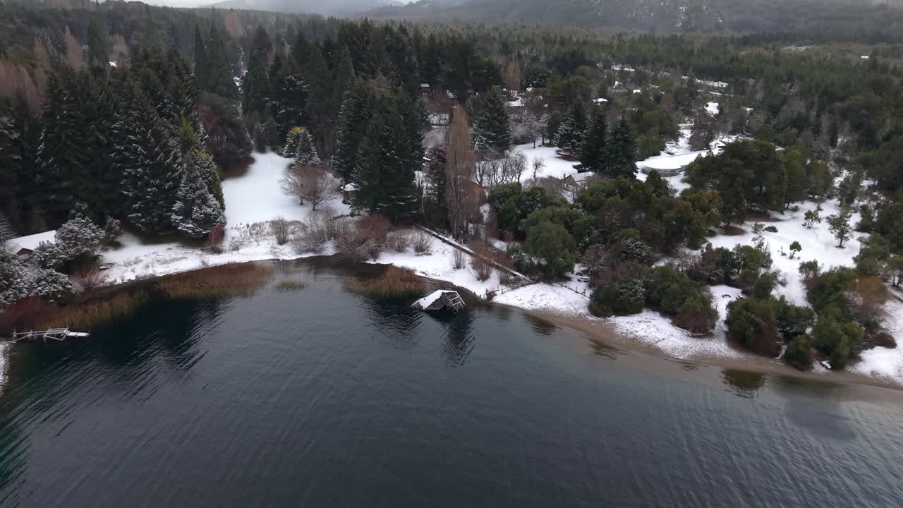 Drone slowly glides over the snowy shoreline of Lake Perito Moreno in Colonia Suiza, Bariloche. Dense pine forest, snowy ground, and cloudy skies frame this iconic Patagonian winter scene