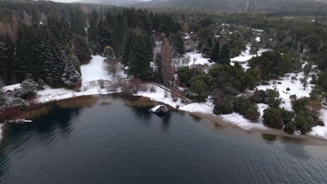 drone slowly glides over the snowy shoreline of lake perito moreno in colonia suiza, bariloche. dense pine forest, snowy ground, and cloudy skies frame this iconic patagonian winter scene